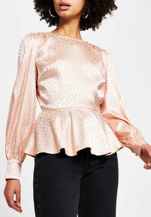 Žena v lesklé broskvové saténové halence s decentním leopardím vzorem, nadýchanými dlouhými rukávy a volánkovým peplum v pase, ve spojení s černými kalhotami.