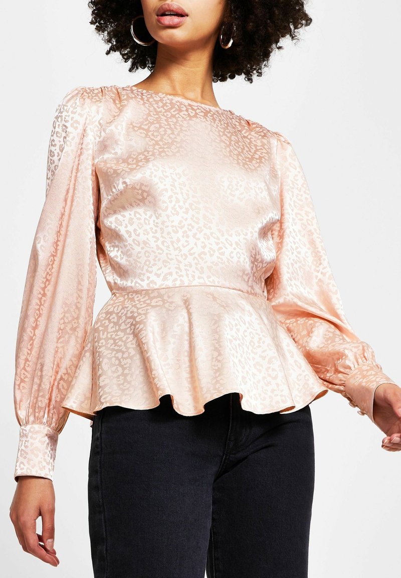 Femme portant un chemisier en satin pêche brillant avec un imprimé léopard subtil, manches longues bouffantes et taille à basques à volants, associé à un pantalon noir.