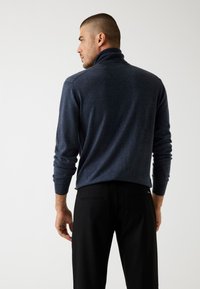 Marineblauer Pullover mit hohem Kragen, gerippten Bündchen und strukturierter Oberfläche, kombiniert mit schwarzen maßgeschneiderten Hosen mit Gesäßtaschen.