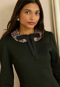 Čierne šaty s golierom so vzorom leopardieho kožúšku a mašľovým detailom. Dlhé rukávy s nariasenými ramenami, hladká textúra. Jemný kontrast voči výstrihu.