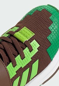 Hnedé textúrované tenisky so zelenými pixelovanými akcentami a logom, s priehľadnou hornou časťou, plochými šnúrkami a bielou podrážkou.