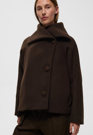 Femme portant un manteau en laine marron foncé avec un grand col et trois boutons recouverts de tissu, debout devant un fond clair uni.