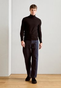 Maglione nero a collo alto, design aderente, abbinato a jeans blu scuri larghi. Le scarpe nere completano l'outfit. Sfondo minimalista con pavimento in legno.