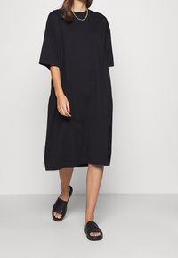 Robe t-shirt noire oversize avec manches courtes, coupe décontractée et longueur genou. Portée avec des sandales à glisser noires, semelle plate texturée.