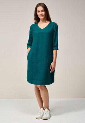Tee-farbendes Leinenkleid mit V-Ausschnitt, Dreiviertelärmeln, seitlichen Taschen und lockerer Passform, kombiniert mit weißen Sneakers vor einem neutralen Hintergrund.