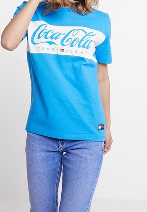 Osoba ubrana w niebieską koszulkę Coca-Cola x Tommy Jeans i jasnoniebieskie dżinsy, stojąca na tle jednolitego białego tła.