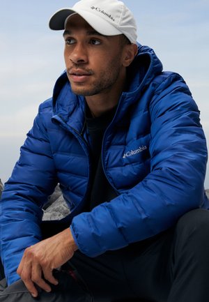 Columbia puffer jacket, blue, quilted, with a zip closure and hood. Worn over a black shirt, paired with a white cap. Textured fabric.