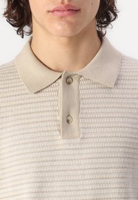 Beige teksturert polo skjorte med en lys beige krage. Har to knapper ved halsen og et subtilt stripet mønster gjennom hele.