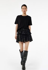 T-shirt noir, jupe à volants noir en sequins superposés, et bottes noires montantes. Tissus lisses avec des embellissements brillants. Silhouette simple et moderne.