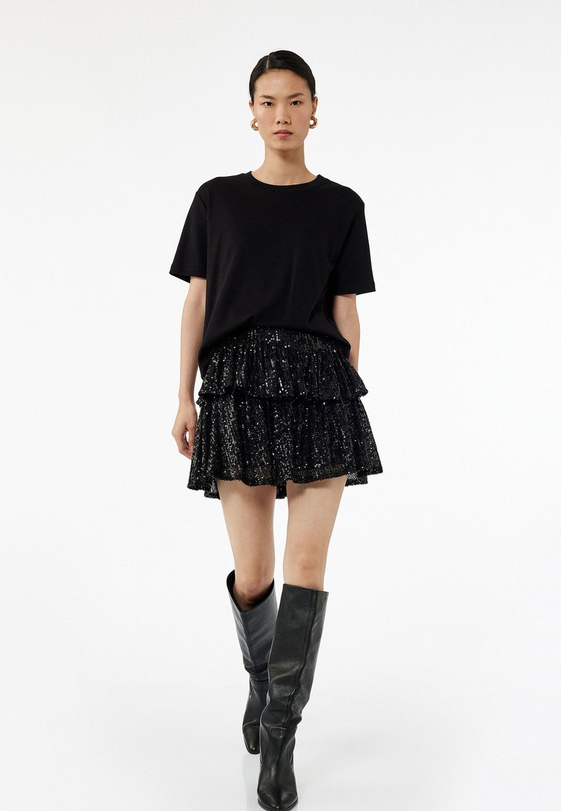 T-shirt noir, jupe à volants noir en sequins superposés, et bottes noires montantes. Tissus lisses avec des embellissements brillants. Silhouette simple et moderne.