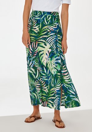 Femme portant une jupe midi verte à imprimé feuilles tropicales avec des boutons sur le côté et des sandales plates marron, debout sur fond blanc.