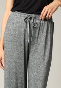 Grijze, metalen, elastische broek met een tailleband met trekkoord. De stof heeft een glans en een zachte textuur. Een hand is zichtbaar die de taille vasthoudt.