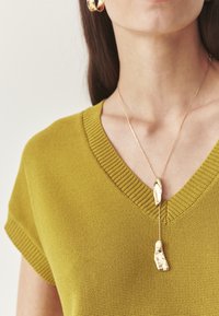 Collier en or avec un pendentif texturé, porté avec un haut en tricot vert à col en V et manches courtes. Texture de tissu lisse et arrière-plan neutre.