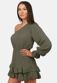 Robe une-épaule vert olive avec un tissu texturé, ourlet à volants, et longue manche ballon, taille cintrée, et ruffles superposés à la jupe.