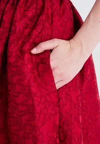 Rød brodert kjole med blomstermønster og teksturert stoff, med sidelomme og rynket midje. 