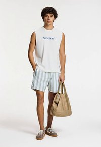 Hvid ærmeløs skjorte med teksten "Suncation", grønne og hvide stribede shorts, grå sneakers og en tan farvet skuldertaske i stof.