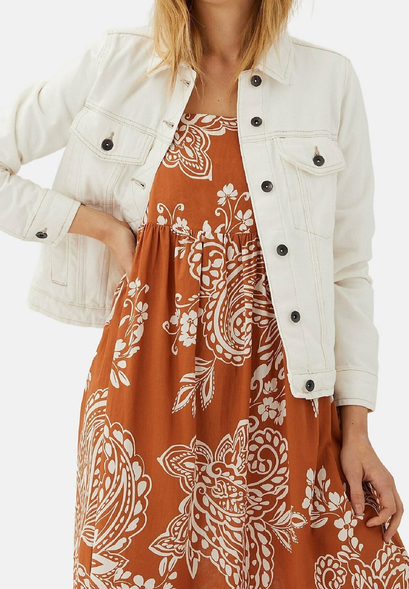 Hvid denimjakke med sorte knapper over en orange kjole med hvide paisley- og blomsterprint. Blødt stof, afslappet pasform og lange ærmer.