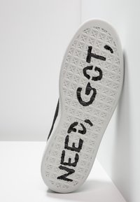 Musta lenkkari tekstuurilla varustetulla päällä, valkoinen kumipohja, jossa on geometrinen kuvio ja musta teksti, joka lukee "NEED GO!" pohjassa.