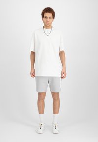 T-shirt blanche en coton oversize, shorts gris avec un logo, et baskets blanches multicolores. Le modèle se tient sur un fond uni.
