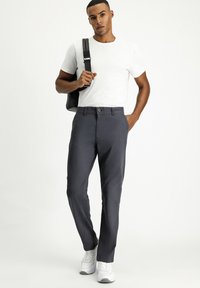 Donkergrijze slim-fit broek met een knoopsluiting en riemlussen, gecombineerd met een aansluitend wit t-shirt en witte sneakers. Zwarte schoudertas.
