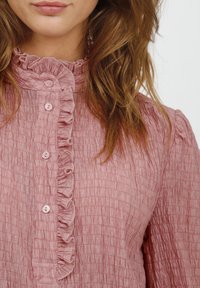Rosa gemusterte Bluse mit einem Rüschkragen und Knopfleiste auf der Vorderseite, ausgestattet mit kleinen Knöpfen und langen Puffärmeln, die einen sanften Schichteffekt erzeugen.