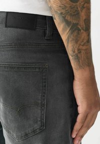 Mørkegrå jeans med en glat tekstur, en baglomme med et subtilt syningdesign og et brandet lædermærke.