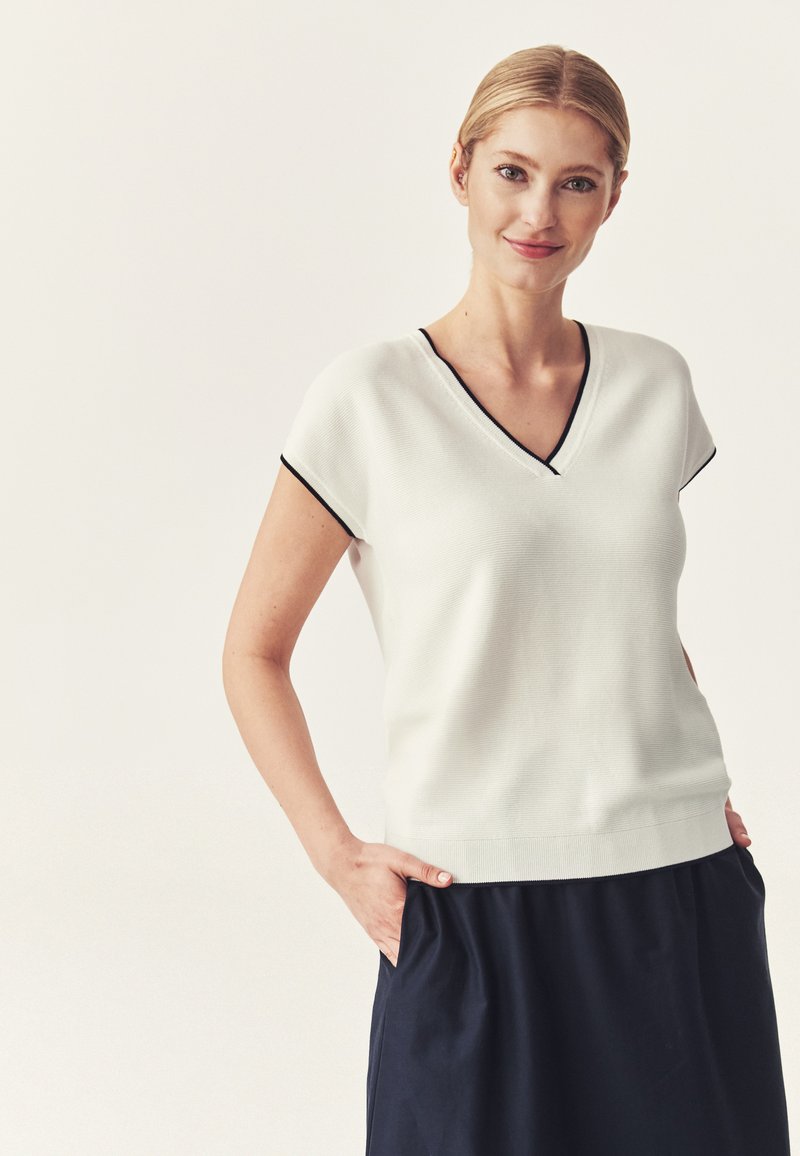 Femme blonde portant un haut blanc à manches courtes en V avec bordure noire et une jupe foncée, souriante, les mains dans les poches.