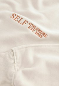 Valge sviiter kangas, millel on tikitud tekst: "SELF ATHLEISURE EST. 2023" helespruuni värvi. Sisaldab ribakäe detaili.