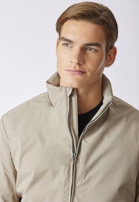 Giacca bomber beige chiaro con colletto alto e chiusura con zip. Texture liscia e design minimalista. Presenta un logo discreto sulla cerniera.