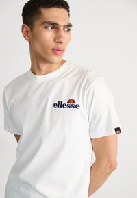 Maglietta di cotone bianca con logo "ellesse" blu navy e stemma colorato, maniche corte e una piccola etichetta con logo sulla manica.