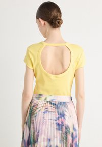 Žlutý top s krátkým rukávem a kruhovým výřezem na zádech, spárovaný s skladanou sukní s barevným květinovým vzorem.