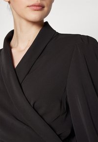 Femme portant un haut noir style cache-cœur avec col châle et détails légèrement bouffants aux épaules sur un fond uni.