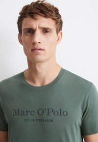 Vihreä puuvillainen T-paita, jossa on pyöreä kaula-aukko ja etupuolella teksti: "Marc O'Polo EST. IN STOCKHOLM" tummansinisellä tekstillä. Sileä rakenne.