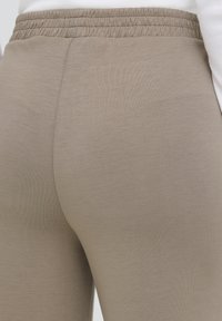 Lähikuva beigenvärisistä elastisista vyötäröhousuista, jotka on pidetty valkoisen paidan päällä, keskittyen takavyötärön ja reiden yläosan alueeseen.