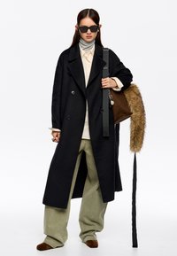 Mujer que lleva un abrigo largo negro, camiseta beige, suéter gris de cuello alto, pantalones holgados oliva, zapatos marrones, gafas de sol negras y un bolso marrón con detalles de piel.