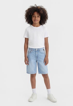 Bambino con capelli ricci che indossa una maglietta bianca semplice, pantaloncini di jeans azzurri, calzini bianchi e scarpe da ginnastica bianche, in piedi contro uno sfondo bianco.