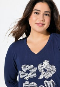 Marinblå långärmad V-ringad topp med ett vitt blommigt grafiskt mönster som har ränder och texturer. Tyget verkar mjukt och ventilerande.
