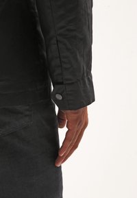 Czarna kurtka z gładkiego materiału, z mankietem na guziki i detalami przy bocznym szwie. Zbliżenie na dłoń spoczywającą obok kurtki.