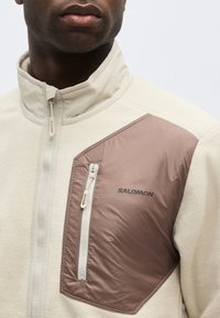 Άνδρας φορούσε μπεζ μπουφάν με ανοιχτό καφέ τσέπη στο στήθος με φερμουάρ και λογότυπο "SALOMON" στην πάνω αριστερή πλευρά.