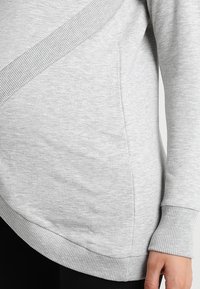 Sweat-shirt gris avec un design croisé, bordure côtelée et texture lisse, ajusté à la taille et aux manches.