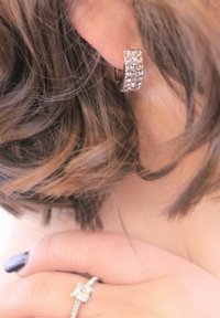 Orecchino a cerchio argentato con design curvo, decorato con molteplici gemme trasparenti. I capelli scuri oscurano parzialmente l'orecchino, con una mano vicina.