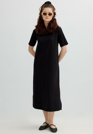 Joven con cabello rizado que lleva un vestido midi negro, gafas de sol beige, bailarinas negras y una coleta negra, de pie con las manos detrás de la espalda.