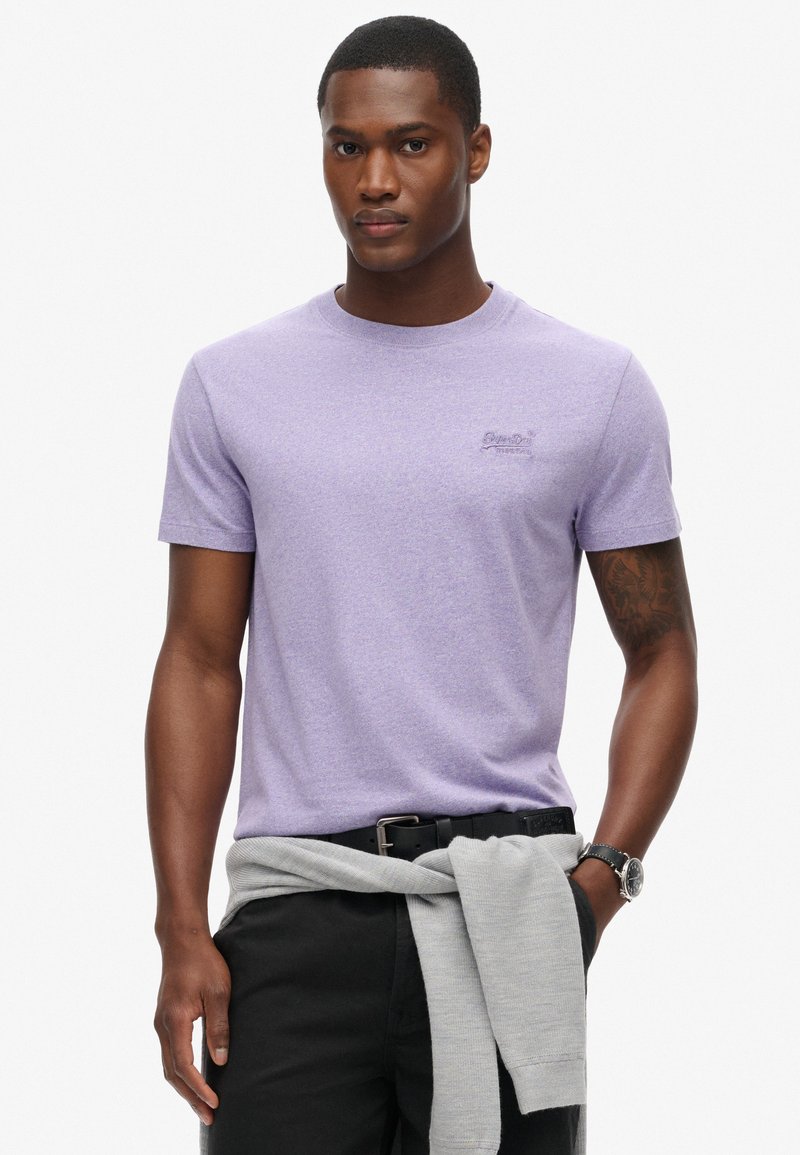 T-shirt girocollo lilla realizzata in tessuto morbido, indossata da un modello. Presenta un piccolo logo ricamato nella parte superiore sinistra del petto.