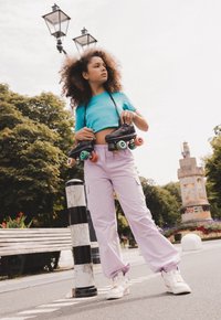 Zwarte rolschaatsen met kleurrijke wielen worden vastgehouden door een persoon in een turkooizen crop top en lichtpaarse cargobroek. Een gestreepte paal staat in de buurt.