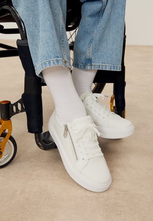 Persona que lleva zapatillas blancas con cordones y cremallera lateral, calcetines blancos y vaqueros azules sentada en una silla de ruedas negra y dorada.