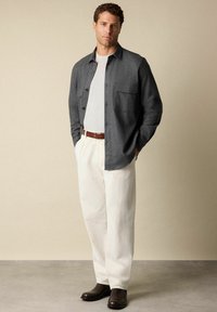 Tmavě šedá košile s knoflíky a kapsou, nošená přes světlou šedou tričko, v kombinaci s bílými kordovými kalhotami a tmavě hnědými botami.