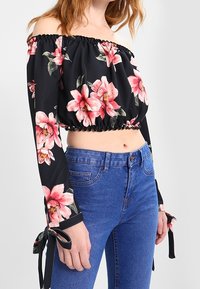 Svart off-shoulder crop top med rosa blommönster, samlad halsringning, långa ärmar med knytning, kombinerad med blå högmidjade jeans.