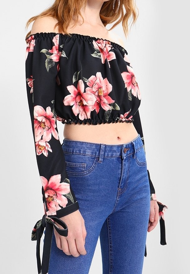 Svart off-shoulder crop top med rosa blommönster, samlad halsringning, långa ärmar med knytning, kombinerad med blå högmidjade jeans.