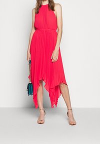 Vestido plisado asimétrico en rosa brillante, con un escote alto y cintura ceñida. Llevado con sandalias con tiras y sosteniendo un bolso color azul verdoso.