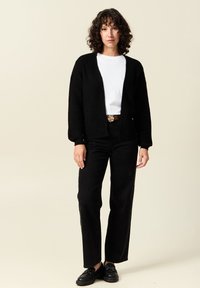 Svart stickad cardigan med avslappnad passform, öppen front och långa ärmar, i kombination med en vit t-shirt, svarta byxor och svarta skor.